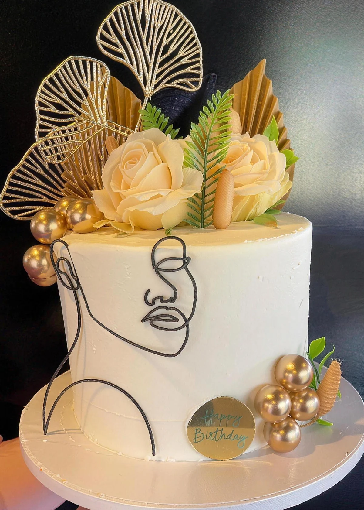 A tall white custom cake with gold leaf fans, cream roses and a line-art portrait